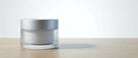 Silver cosmetic jar with frosted glass body and metallic cap on wooden surface against white background for skincare product packaging.