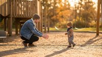 Young father crouching with open arms encouraging toddler son to walk on playground during golden hour sunset creating warm family bonding moment outdoors.