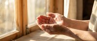 Elderly hands cupped in prayer or meditation gesture by warm sunlit window, expressing faith, hope, spirituality and peaceful contemplation in golden light.
