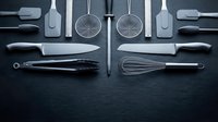 Professional kitchen utensils and tools arranged symmetrically on dark slate surface. Modern cooking equipment for culinary preparation and food service.