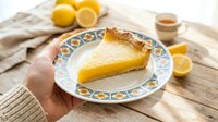 Fresh lemon tart slice on decorative plate held by hand, golden custard filling with flaky pastry crust, citrus dessert ready for serving on rustic wooden table.