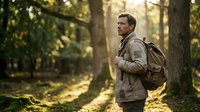 Middle-aged Caucasian male hiker wearing brown jacket and backpack, walking through sunlit forest path surrounded by tall trees and green foliage during daytime outdoor adventure.