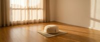 Meditation cushion on yoga mat in sunlit room with wooden floors and sheer curtains creating peaceful mindfulness space for relaxation and wellness practice.