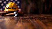 Yellow construction hard hat with Labor Day text against blurred American flag background on rustic wooden surface, selective focus composition.