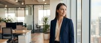 Young professional woman with dark hair and South Asian ethnicity wearing a navy blazer and beige blouse, standing by floor-to-ceiling windows in a modern office environment with city views, suitable for corporate, business, and professional themes.