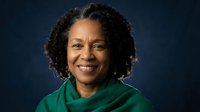Professional African American businesswoman with curly hair smiling confidently in green blazer against dark blue background for corporate headshots.