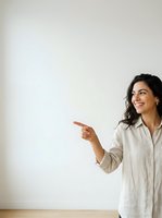 Woman with long dark hair and olive complexion wearing a beige linen shirt, smiling while pointing to the left against a white wall. Suitable for business, education, and presentation materials.