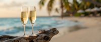 Two champagne flutes filled with sparkling wine on driftwood at tropical beach during golden hour sunset with palm trees and turquoise ocean in background.
