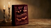 Vintage Halloween book with ornate cover featuring bats and jack-o-lanterns, displayed with flickering candles on rustic wooden surface for spooky seasonal decor.
