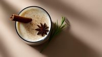 Creamy eggnog cocktail with cinnamon stick, star anise, and pine garnish on neutral background with soft shadows.
