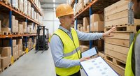 Warehouse worker in safety vest and hard hat checking inventory with clipboard in modern distribution center with stacked boxes on shelves.