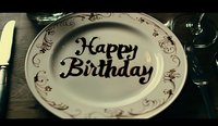 Elegant white plate with ornate gold trim and Happy Birthday text in black calligraphy script, photographed on dark wooden table with moody lighting.
