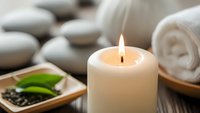 Spa wellness scene with burning candle, rolled towels, smooth stones and green leaves creating peaceful relaxation atmosphere for massage therapy treatments.