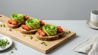 Gourmet open-faced sandwiches topped with creamy avocado roses, roasted tomatoes and fresh herbs on toasted bread slices arranged on wooden board for catering and food service.