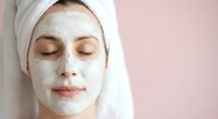 Young woman with closed eyes enjoying white clay facial mask treatment wearing towel on head against pink background for skincare and spa wellness routine.