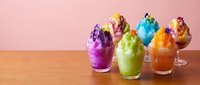 Colorful shaved ice desserts in glass cups with syrup toppings arranged on wooden surface against pink background, perfect for food and beverage marketing.