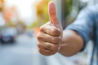 Close-up of Asian male hand showing thumbs up gesture against blurred urban background, expressing positivity, agreement and satisfaction.