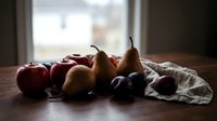 Fresh apples, pears, and plums arranged on a rustic wooden table near a window, creating a moody autumn still life scene.