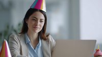Young Caucasian businesswoman in party hat smiling at laptop during office celebration, expressing joy and satisfaction at work.