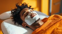 Young African American woman relaxing with hydrating face mask during spa treatment, lying on massage table covered with bright orange towel in warm sunlight.