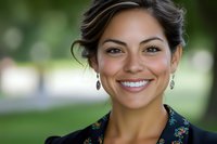 Young woman with dark hair and olive complexion smiling outdoors in park setting, wearing dark patterned shirt, professional headshot for business, corporate, or personal branding use.