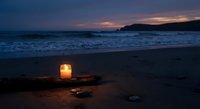 Glowing lantern on sandy beach at twilight with ocean waves and mountain silhouette creating peaceful coastal atmosphere for relaxation and meditation.