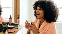 Happy African American woman with curly hair applying skincare products in bright bathroom mirror reflection morning routine self-care wellness beauty ritual.