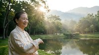 Woman in casual shirt standing by riverside at sunrise, enjoying peaceful nature landscape with mountains and trees. Suitable for wellness, lifestyle, and travel content.