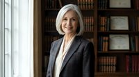 Professional mature Caucasian businesswoman with blonde bob haircut smiling confidently in elegant dark suit standing in library office environment.