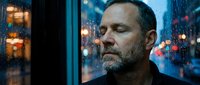 Middle-aged man with beard gazing through rainy window at night cityscape with bokeh lights, contemplative mood, urban setting.