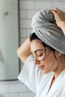 Young woman wrapping wet hair in gray towel after shower in modern bathroom. Hair care routine, personal hygiene and wellness concept.