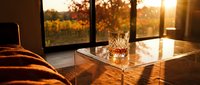 Glass of whiskey on table with autumn landscape visible through windows during golden hour sunset.