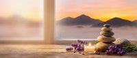 Zen meditation setup with balanced stones, lavender flowers and candle on wooden windowsill overlooking misty mountain landscape at golden sunrise.