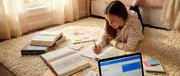 Young female student of South Asian descent studying on bedroom floor with laptop, notebooks, and educational materials for homework, online learning, and academic preparation.