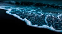 Dramatic close-up of ocean foam and waves in dark blue tones creating abstract patterns on black sandy beach, moody seascape with white seafoam texture.
