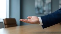 Businessman in dark suit extending open palm gesture on wooden table, offering help or presenting proposal in professional office setting.