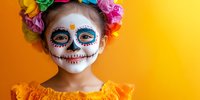 Young girl with traditional sugar skull face paint and colorful flower crown wearing orange dress against vibrant yellow background celebrating Day of Dead festival.