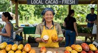 Smiling Asian woman farmer selling fresh local cacao pods at outdoor market stand. Sustainable agriculture and organic farming concept.