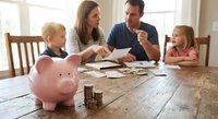 Caucasian family with two children learning about money management and savings at wooden dining table with pink piggy bank and coins in bright home interior.