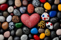 Colorful collection of painted stones with red heart shape in center surrounded by pebbles decorated with dots, patterns and various designs on dark background.