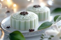 Traditional Asian snow skin mooncakes with delicate floral pattern on white plate, garnished with green leaves and bokeh lights in background.