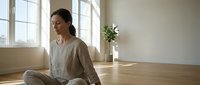 Young Asian woman practicing mindful meditation in bright minimalist room with wooden floors and natural light for wellness and mental health.