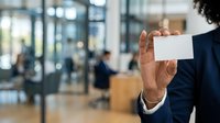 Young male professional in dark business suit holding blank white business card in modern office environment with blurred colleagues in background.