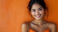 Young Asian woman with bright genuine smile and natural makeup against vibrant orange wall background, wearing spaghetti strap top. Casual lifestyle portrait.