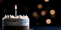 Slice of chocolate and vanilla cake with lit blue birthday candle against dark background with soft bokeh lights, celebration mood and copy space.