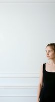 Young woman with light brown hair, fair complexion, wearing black sleeveless dress and pearl necklace, standing against white wall with copy space for design projects.