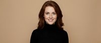 Young Caucasian woman with wavy brown hair wearing black turtleneck sweater smiling at camera against neutral beige background for professional headshot.