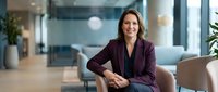 Confident Caucasian businesswoman in burgundy blazer sitting in modern office lobby, smiling warmly, conveying leadership and professionalism.