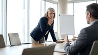Mature woman with blonde hair and light skin tone, wearing dark blazer, leaning forward over conference table during business meeting with male colleague in modern office with large windows.
