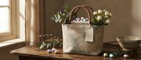 Natural linen Easter basket with colorful eggs and spring flowers on rustic wooden table by sunny window for holiday celebration and seasonal home decor.